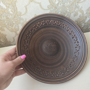 May include: A brown ceramic plate with a decorative floral pattern around the rim. The plate has a slightly raised edge and a central recessed area. The plate is held by a hand with pink nail polish.
