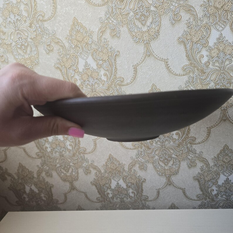 May include: A dark brown, shallow ceramic bowl with a slightly curved rim. The bowl is held in a hand, showcasing its smooth, matte finish. The background features a patterned wallpaper.