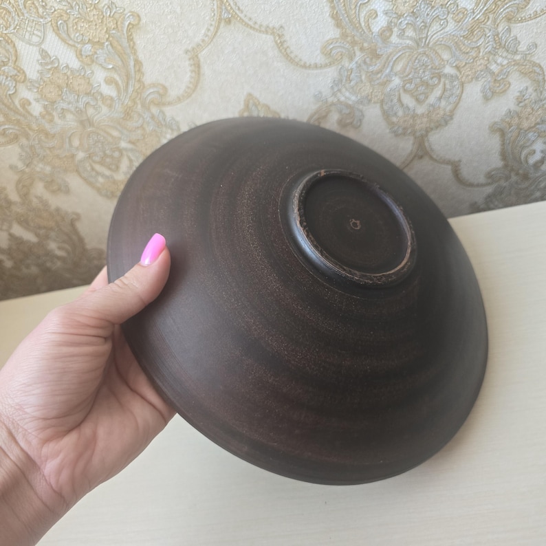 May include: A dark brown ceramic bowl with a circular base. The bowl has a smooth, matte finish and a slightly textured surface. The bowl is held in a hand with pink nail polish.