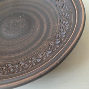 May include: A handmade, dark brown ceramic plate with a decorative floral pattern around the rim. The plate has a rustic, earthy aesthetic, with visible texture and a slightly uneven surface. The plate's rim is a lighter brown color.