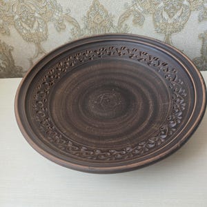 May include: A round, brown ceramic plate with a decorative floral border. The plate has a wide rim and a slightly raised center. The plate is made of a dark brown clay and has a rustic, handmade appearance.