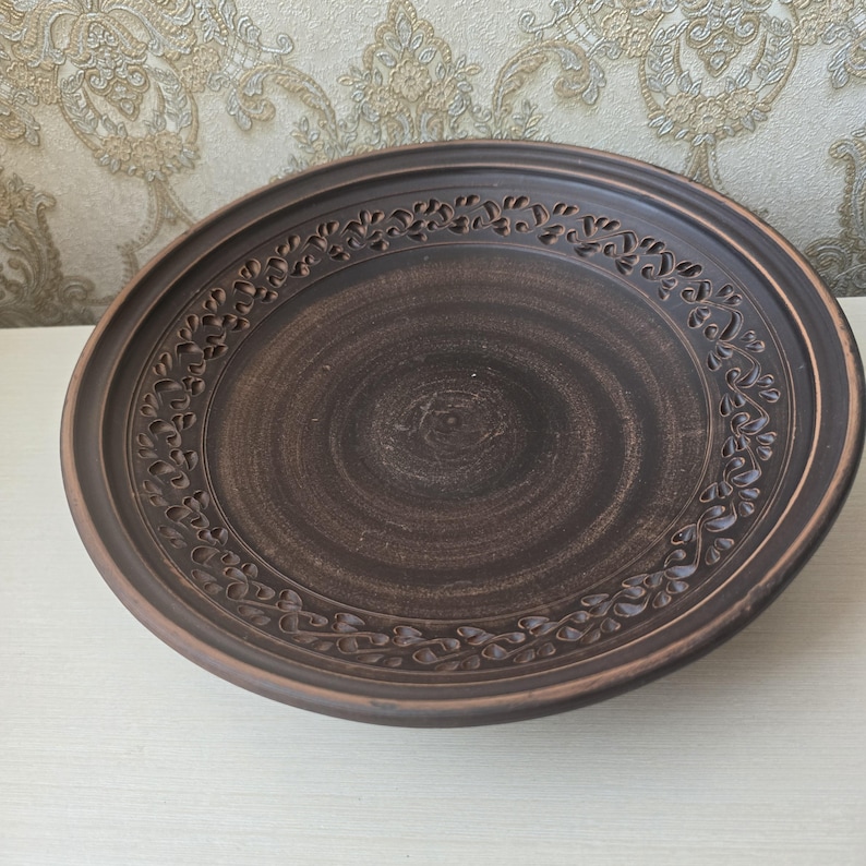 May include: A round, brown ceramic plate with a decorative floral border. The plate has a wide rim and a slightly raised center. The plate is made of a dark brown clay and has a rustic, handmade appearance.