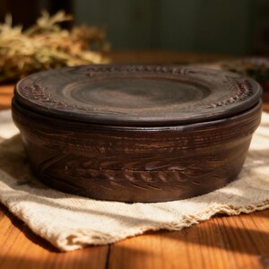 May include: A handcrafted, dark brown ceramic bowl with a lid. The bowl and lid feature embossed wheat stalk designs. The bowl is sitting on a beige cloth, with a wooden table in the background.