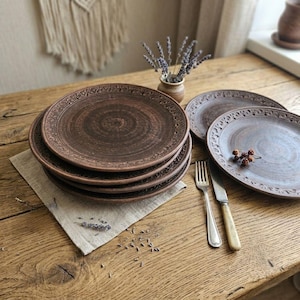 May include: A stack of six brown ceramic plates with a decorative border and a spiral design in the center. The plates are on a beige linen napkin on a wooden table. A fork and knife are next to the plates.