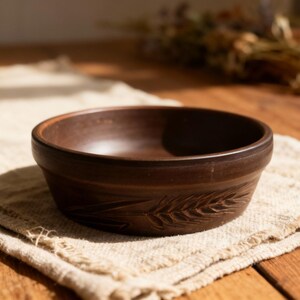 Op de afbeelding: Een bruine keramische kom met een tarwe-aar ontwerp in de zijkant gesneden. De kom staat op een beige linnen doek. De kom is ongeveer 5 cm hoog en 15 cm in diameter. De achtergrond is een houten tafel.