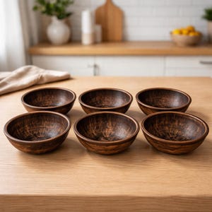 Pode incluir: Seis pequenas tigelas redondas de madeira com acabamento castanho escuro estão dispostas sobre uma superfície de madeira castanha clara. As tigelas têm um interior texturizado e um design simples e rústico, adequadas para servir lanches ou porções pequenas.