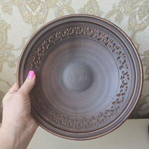 May include: A brown ceramic plate with a decorative floral border. The plate has a slightly raised rim and a central depression. The plate is held by a hand with pink nail polish.