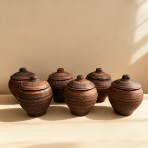 Pode incluir: Um conjunto de seis potes de cerâmica marrom com tampas. Cada pote, de formato arredondado, é decorado com padrões gravados e um pequeno botão na tampa. Estão dispostos sobre uma superfície neutra.