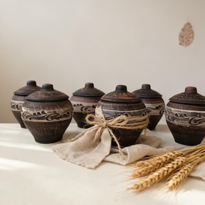 Może przedstawiać: Kolekcja sześciu ręcznie robionych, ciemnobrązowych ceramicznych słoików z pokrywkami. Każdy słoik ma dekoracyjny biały wzór fal w środku. Jeden słoik jest ozdobiony kokardą ze sznurka i spoczywa na beżowej tkaninie, z kłosami pszenicy na pierwszym planie.