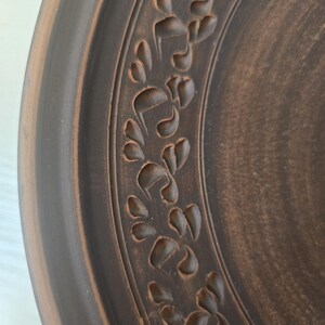 May include: Close-up of a brown ceramic plate with a decorative floral pattern carved around the rim. The plate has a rustic, handmade appearance with variations in the brown color.