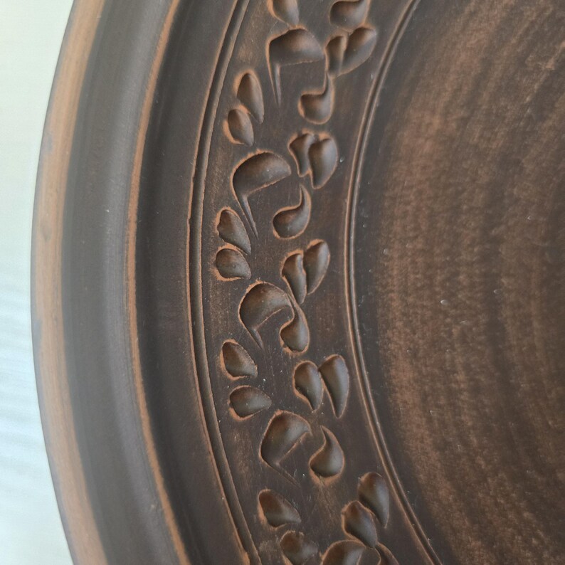 May include: Close-up of a brown ceramic plate with a decorative floral pattern carved around the rim. The plate has a rustic, handmade appearance with variations in the brown color.