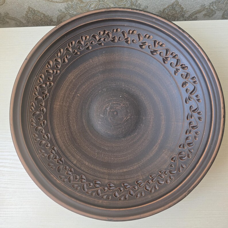 May include: A brown ceramic serving bowl with a decorative floral pattern around the rim. The bowl has a wide, shallow design and a dark, earthy color, suitable for serving food.