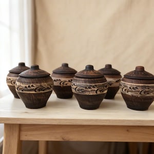 Könnte beinhalten: Sechs handgefertigte, dunkelbraune Keramikgläser mit Deckeln. Jedes Glas hat ein geschnitztes wellenförmiges Design in der Mitte und einen abgerundeten Deckel. Die Gläser stehen auf einem hellen Holztisch und unterstreichen ihren rustikalen Charme.