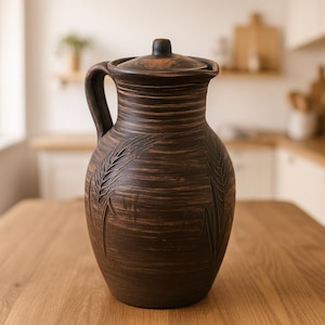 Può includere: Una brocca in ceramica marrone con coperchio e manico, con spighe di grano incise. La brocca ha un aspetto rustico e artigianale, con linee orizzontali sul corpo. È appoggiata su una superficie di legno.