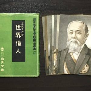 以下が含まれることがあります： 白ひげの立派な男性の肖像画が描かれた、日本のヴィンテージ絵葉書のコレクション。緑色の本が隣にあり、日本語のテキストが書かれています。本の表紙には白黒の文字が書かれています。