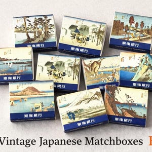 以下が含まれることがあります： ヴィンテージ日本のマッチ箱のコレクション。各箱には、青、緑、茶色の異なる風景画が描かれています。箱には日本語のテキストが入った青い縁があります。「Vintage Japanese Matchboxes Etsy」というテキストが下にあります。