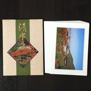 以下が含まれることがあります： 京都の清水寺の風景写真が写された絵葉書のセット。絵葉書は、清水寺のダイヤモンド型の画像と日本語のテキストが書かれた装飾的な箱の隣に積み重ねられています。箱は、花の模様が入ったベージュ色の背景です。