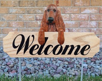 May include: A wooden sign with the word "Welcome" in black script, held by a carved, reddish-brown dog. The sign is mounted on metal stakes and placed in a garden with grass and pebbles. A brick wall is in the background.