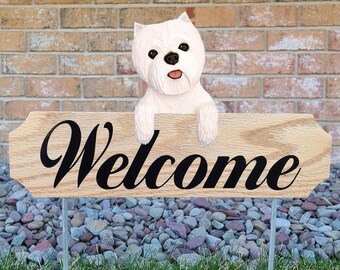 May include: A wooden welcome sign with a white West Highland White Terrier dog peeking over the top. The sign is on a metal stake and is in front of a brick wall.