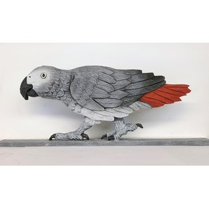 May include: A carved wooden parrot sculpture in shades of gray with a black beak and a vibrant red tail. The bird is depicted in a walking pose, mounted on a gray rectangular base. The artwork is set against a white background.