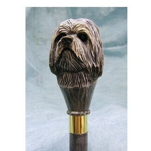 May include: A brown wooden walking stick with a carved Shih Tzu dog head handle. The handle is detailed with fur and facial features. The stick has a gold metal band around the shaft.