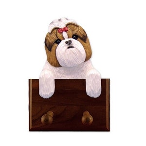 Puede incluir: Un perchero de madera con dos ganchos presenta una figurita esculpida de un perro Shih Tzu marrón y blanco con un lazo rojo. El perro está mirando por encima de la parte superior del perchero.