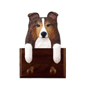 Puede incluir: Un gancho de madera para la correa de un perro con un perro Shetland tallado en madera, con pelo marrón y blanco. El perro asoma por encima del gancho, que tiene dos ganchos en la parte inferior.
