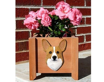 May include: A wooden planter box with a carved corgi dog head on the front. The planter is filled with pink flowers and is sitting on a brick surface.