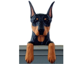May include: A wooden figurine of a black and tan miniature Doberman Pinscher dog peeking over a fence. The dog has a serious expression and its paws are resting on the top of the fence.