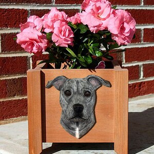 May include: A wooden planter box with a gray pit bull dog face carved into the front. The planter is filled with pink flowers.