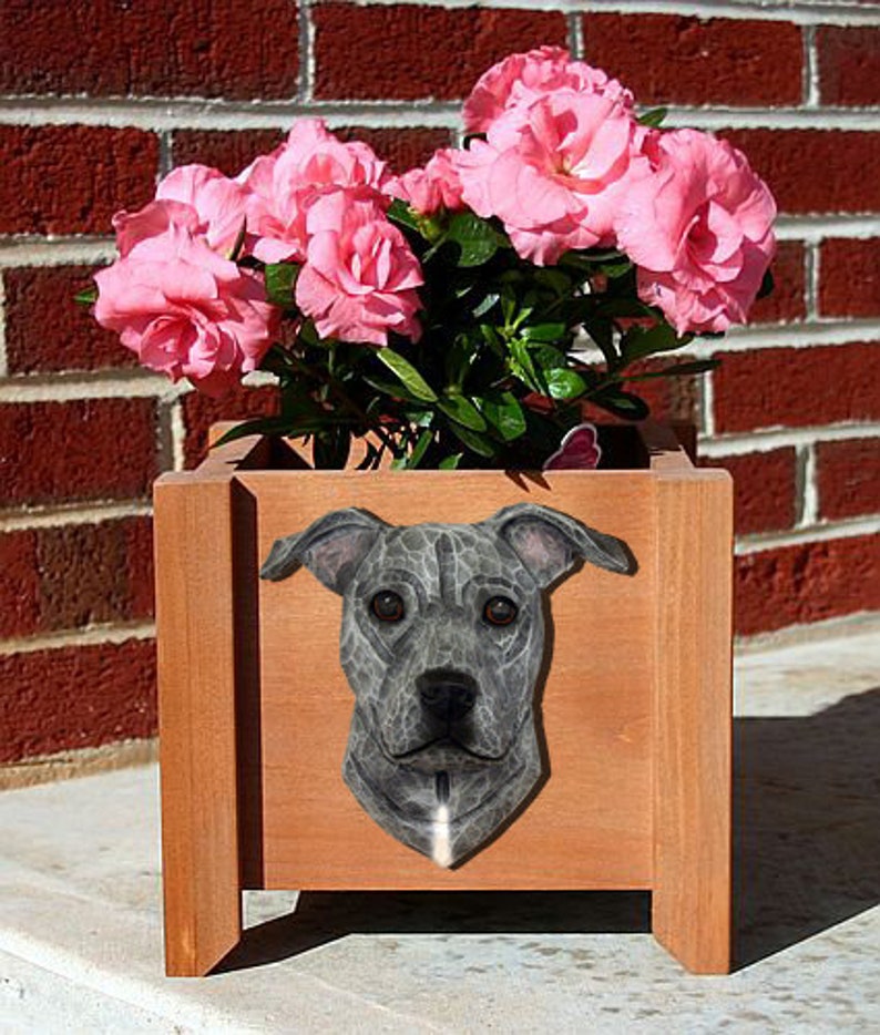 May include: A wooden planter box with a gray pit bull dog face carved into the front. The planter is filled with pink flowers.