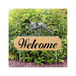 May include: A garden welcome sign with a bronze dog sculpture above a wooden plaque. The plaque displays the word "Welcome" in a stylish script. The sign is supported by metal stakes and set against a backdrop of green foliage.