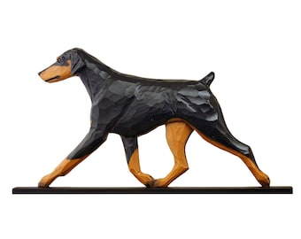 May include: A wooden sculpture of a Doberman Pinscher dog in a walking pose. The dog is black with tan markings on its snout, legs, and paws. The sculpture is mounted on a black base.