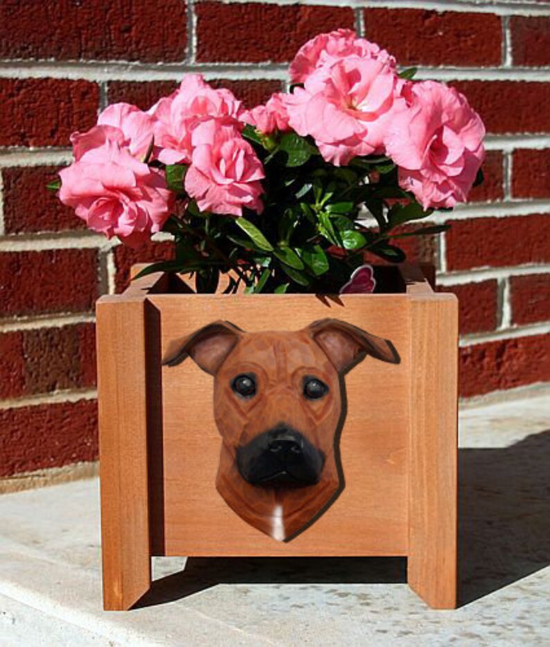May include: A wooden planter box with a brown dog's head carved into the front. The planter is filled with pink flowers.