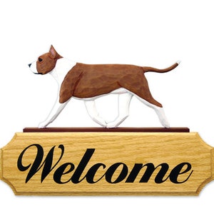 May include: A wooden welcome sign with a carved brown and white American Bulldog walking across the top. The sign is made of light brown wood and has the word "Welcome" in black lettering.