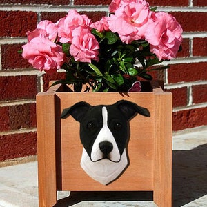 May include: A wooden planter box with a black and white dog sculpture attached to the front. The planter is filled with pink flowers.