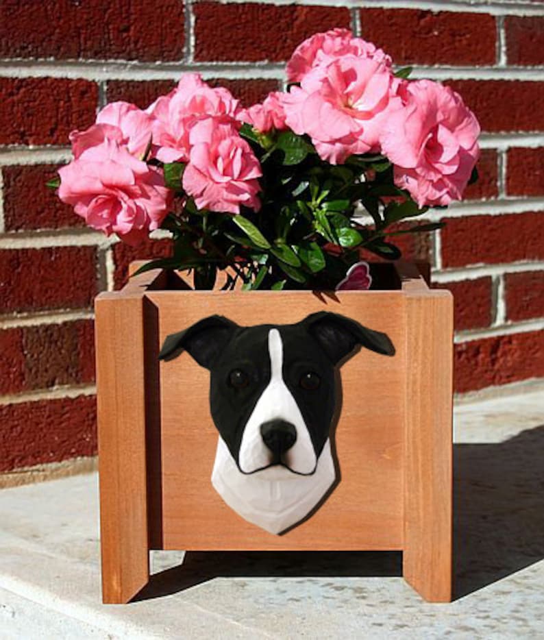 May include: A wooden planter box with a black and white dog sculpture attached to the front. The planter is filled with pink flowers.