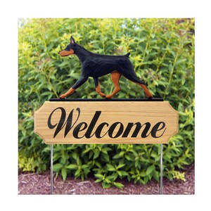 May include: A wooden sign with the word "Welcome" in black script. A carved black and tan Doberman dog figure is mounted above the sign. The sign is on metal stakes, set against a backdrop of green foliage.