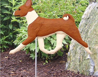 May include: A hand-carved wooden garden stake shaped like a Basenji dog. The figure features a reddish-brown coat with white markings on the chest, muzzle, and legs, and a curled tail, mounted on a thin metal rod for ground placement.