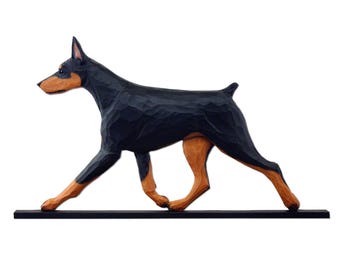 May include: A wooden sculpture of a Doberman Pinscher dog in a walking pose. The dog is black with tan markings on its legs, chest, and face. The sculpture is mounted on a black base.