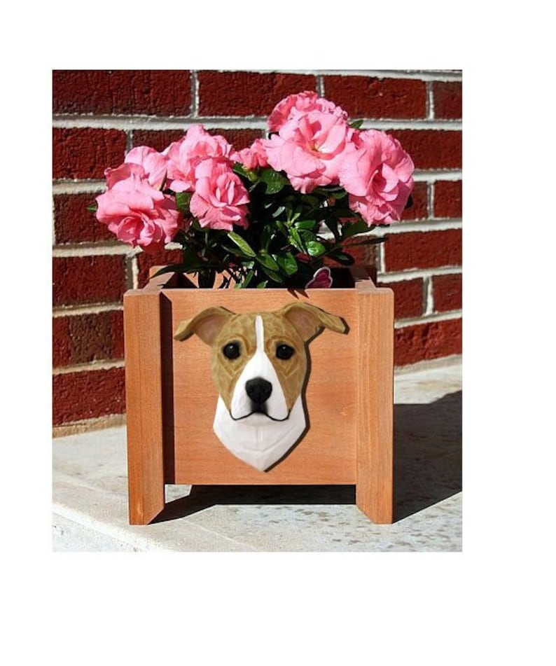 May include: A wooden planter box with a brown and white dog head sculpture on the front. The planter is filled with pink flowers.