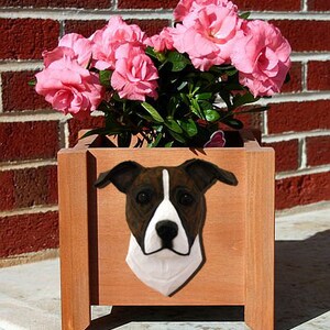 May include: A wooden planter box with a brown finish features a white and brown dog head sculpture. The planter is filled with pink flowers.
