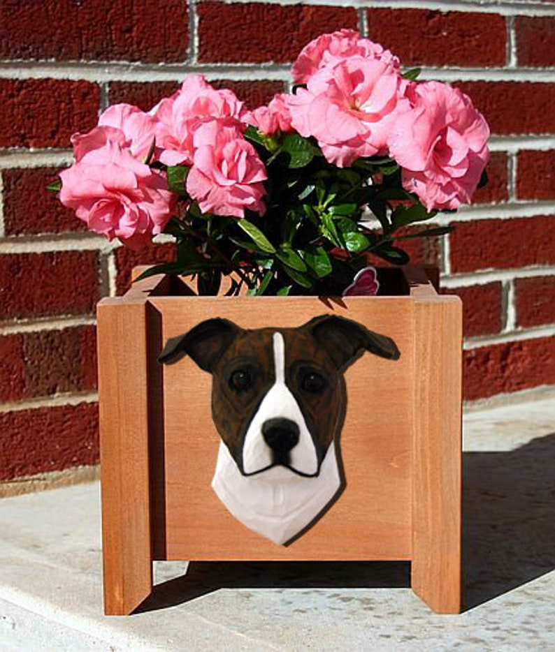 May include: A wooden planter box with a brown finish features a white and brown dog head sculpture. The planter is filled with pink flowers.