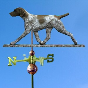 May include: A metal weather vane featuring a dog silhouette with brown and white markings. The dog is mounted above a directional compass with gold and green accents, set against a blue sky background.