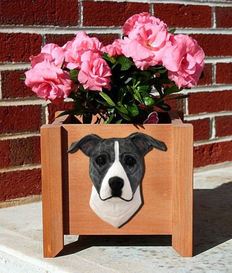 May include: A wooden planter box with a black and white dog head sculpture on the front. The planter is filled with pink flowers.