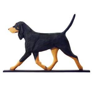 Op de afbeelding: Houten beeldhouwwerk van een Bloodhound-hond in een wandelhouding. De hond is voornamelijk zwart met bruine accenten op de poten en snuit. Het beeldhouwwerk is gemonteerd op een zwarte basis.
