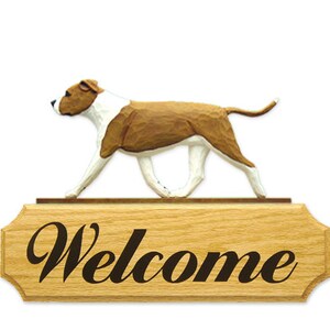 May include: Wooden welcome sign with a carved brown and white Staffordshire Bull Terrier dog figurine on top. The sign reads "Welcome" in a black cursive font.