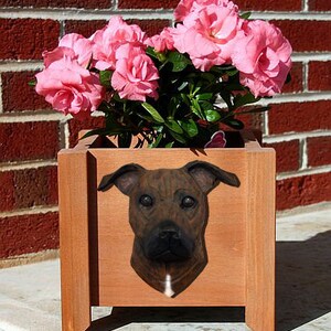 May include: A brown wooden planter with a brown and black dog head sculpture on the front. The planter is filled with pink flowers.