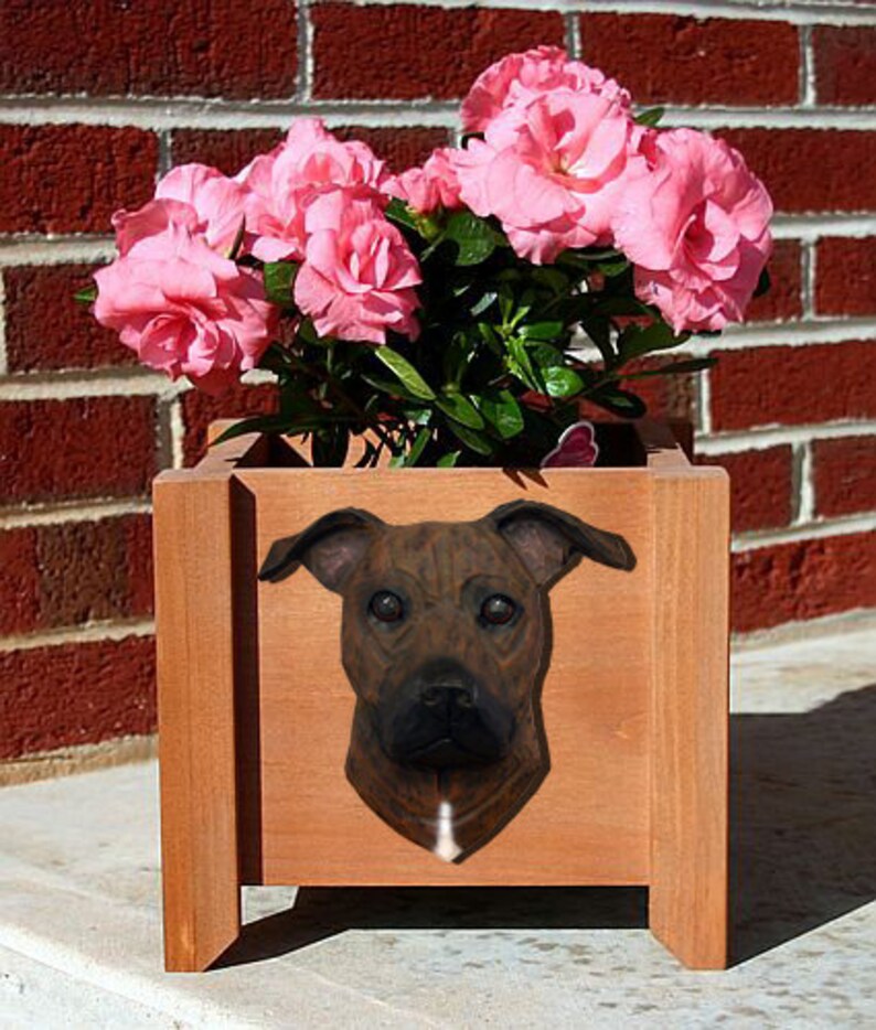 May include: A brown wooden planter with a brown and black dog head sculpture on the front. The planter is filled with pink flowers.