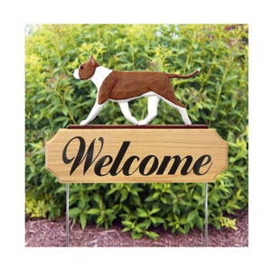 May include: A wooden yard sign featuring a brown and white dog sculpture above the word "Welcome" in black script. The sign is mounted on metal stakes, set against a backdrop of green foliage.